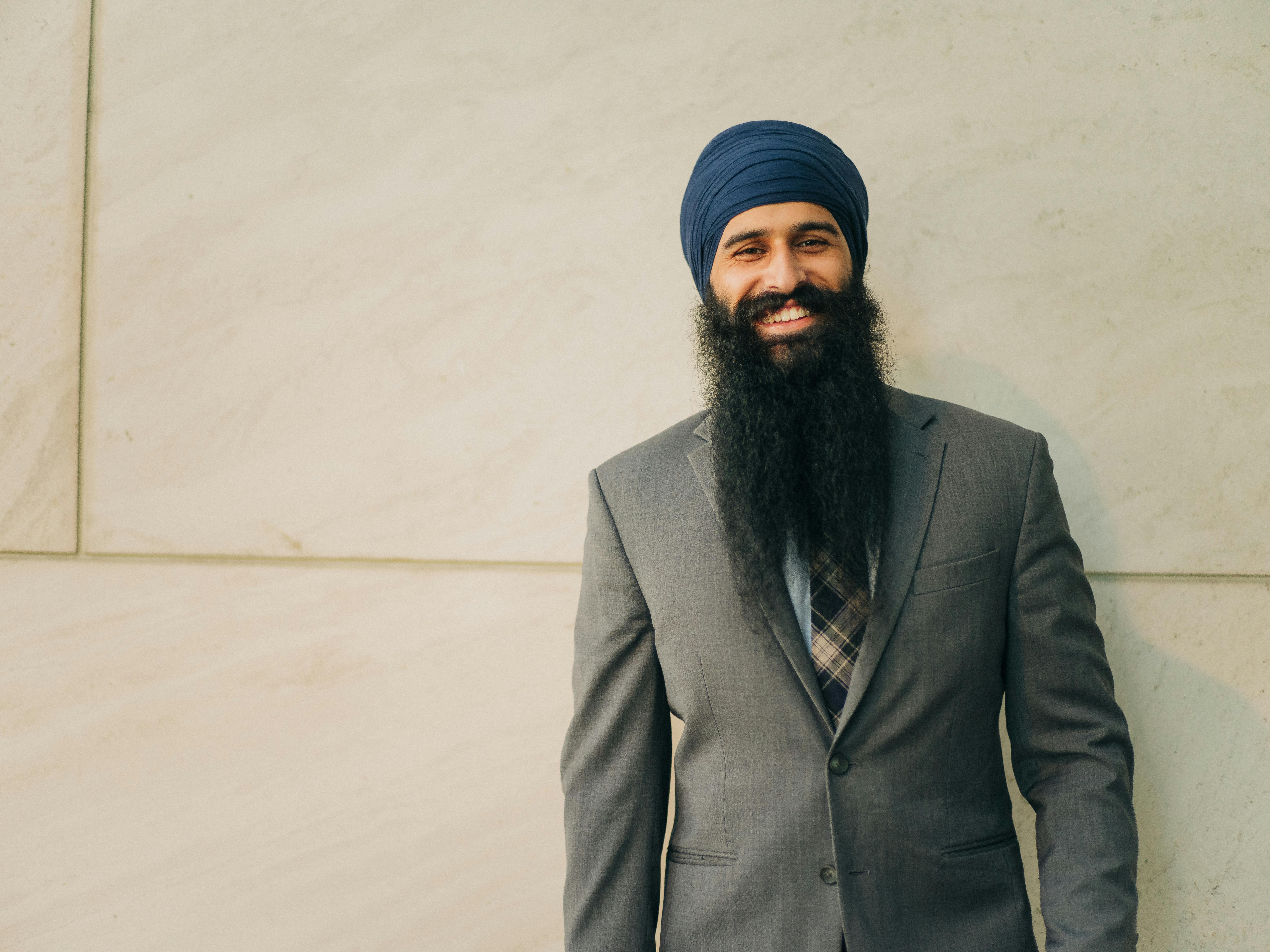 Sikh businessman in full suit and turban smiling confidently against a cream wall.